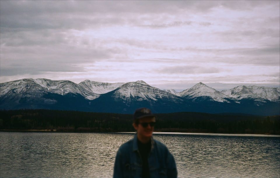 A smiling person in a denim jacket, cap and sunglasses, out of focus, stands in front of a lake and snow-capped mountains under a cloudy sky.