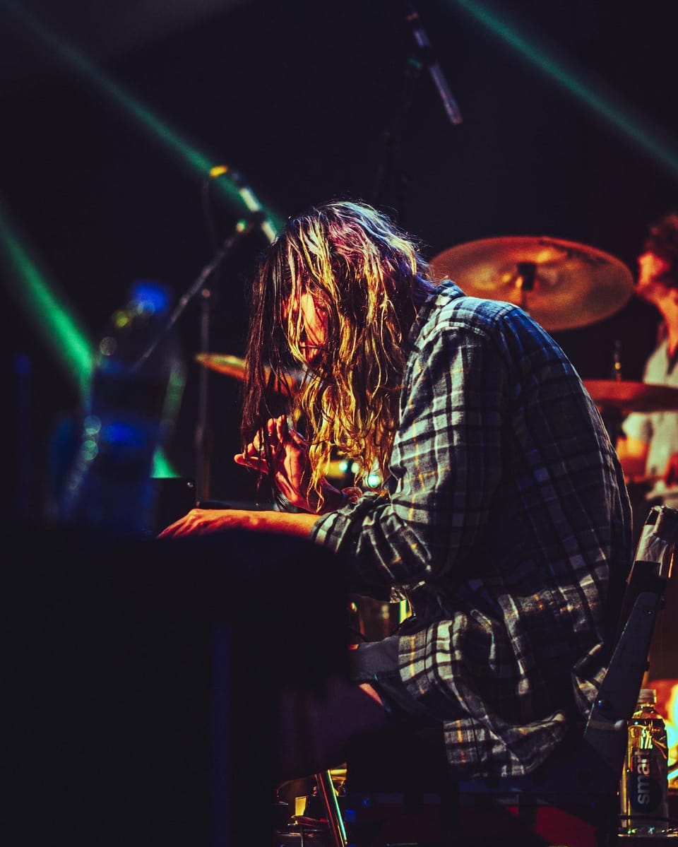 A long-haired musician in a plaid shirt leans over a keyboard in green and blue stage light.