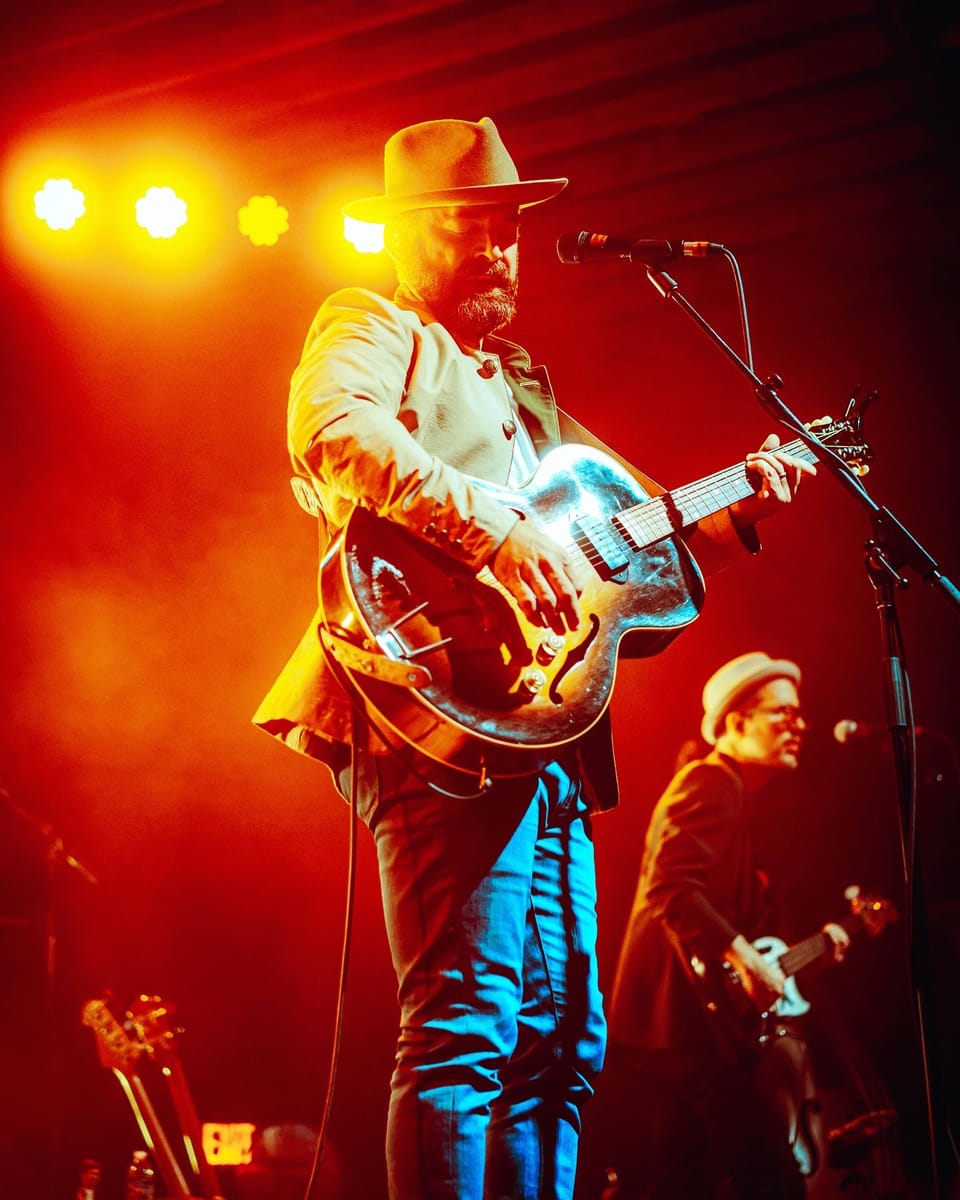 Drew Holcomb. A singer in a cowboy hat plays a resonator guitar under hot red and orange lights.