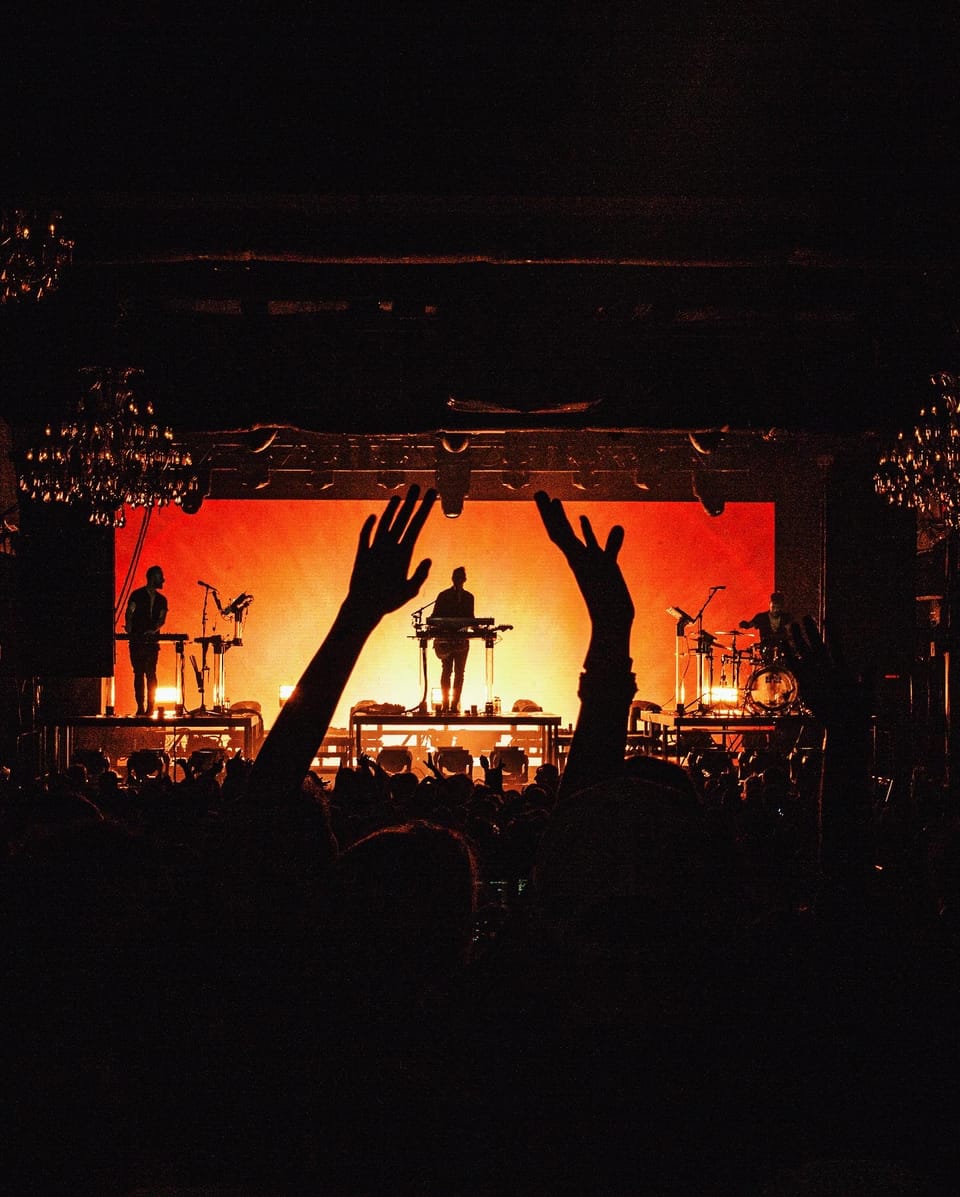 Rufus Du Sol. Silhouetted hands reach up toward a stage glowing orange, seen from the crowd.