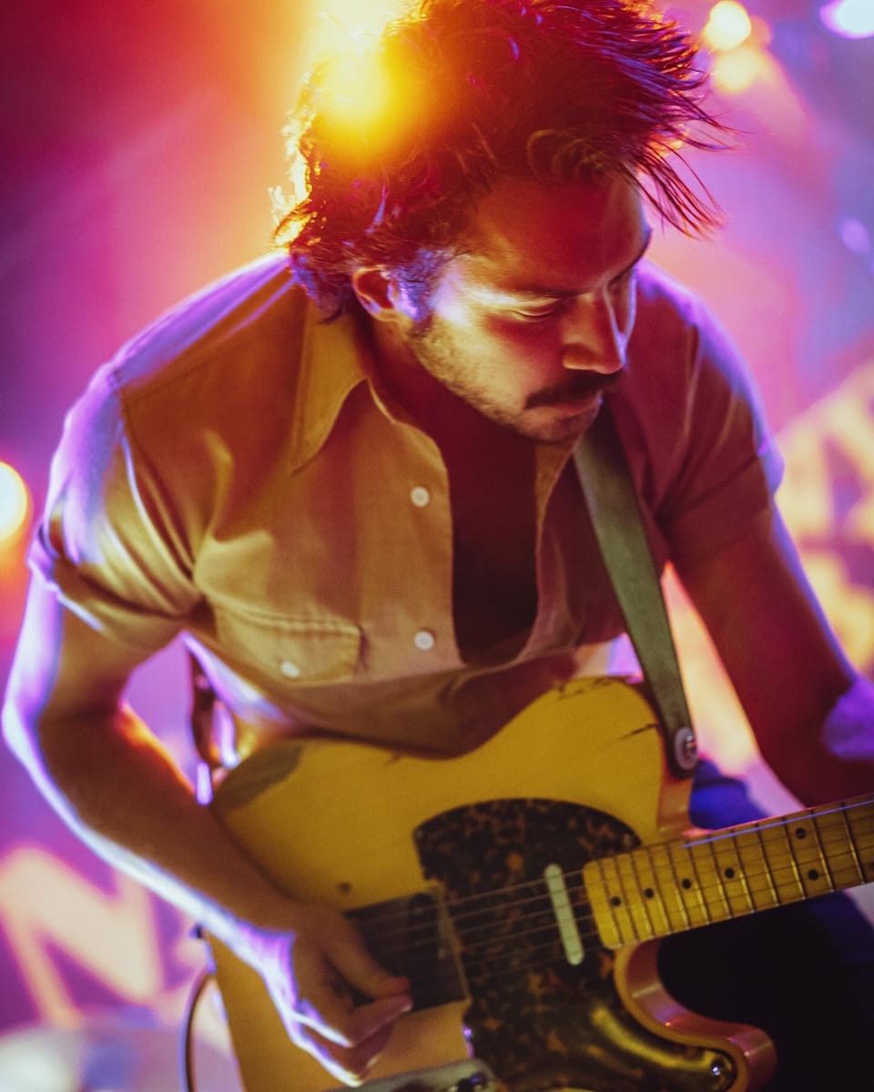 Mo Lowda and the Humble. A close, tilted portrait of a guitarist in a yellow shirt playing a Telecaster in purple light.