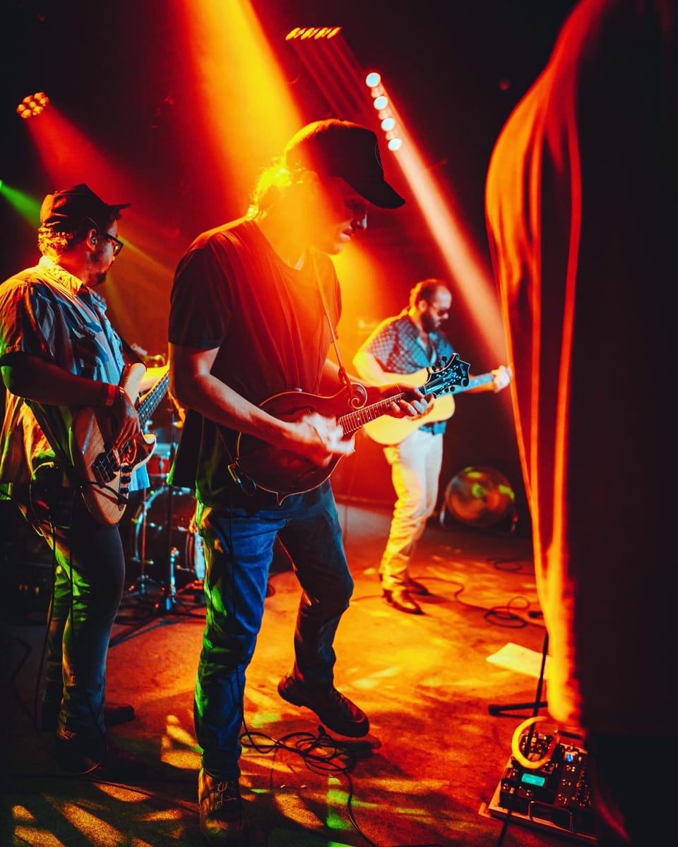 Town Mountain. Three musicians in caps and jackets play on a stage lit by orange beams, beside a red curtain.