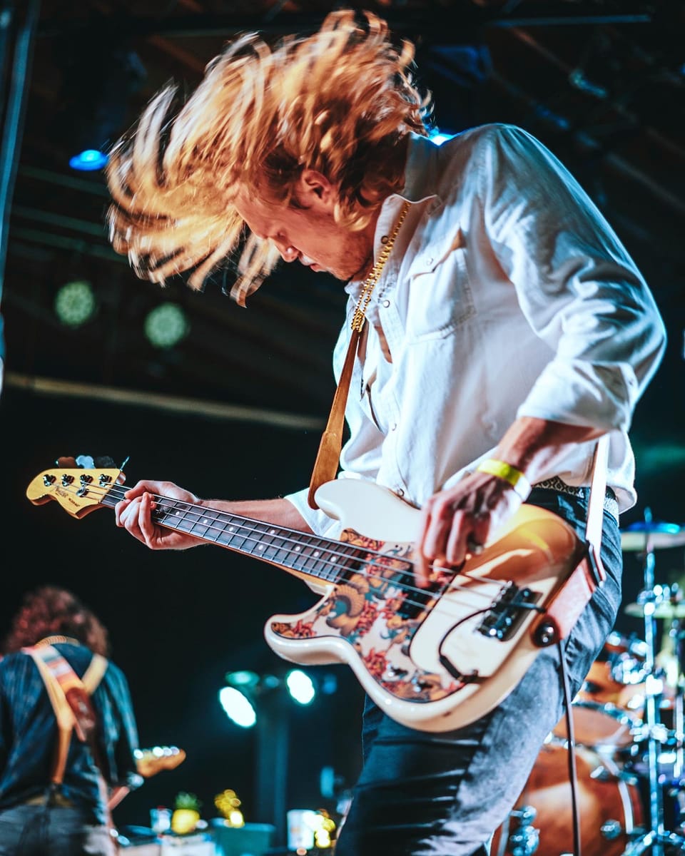 The Stews. A bassist in a white shirt, blond hair flying, plays a floral-patterned bass.