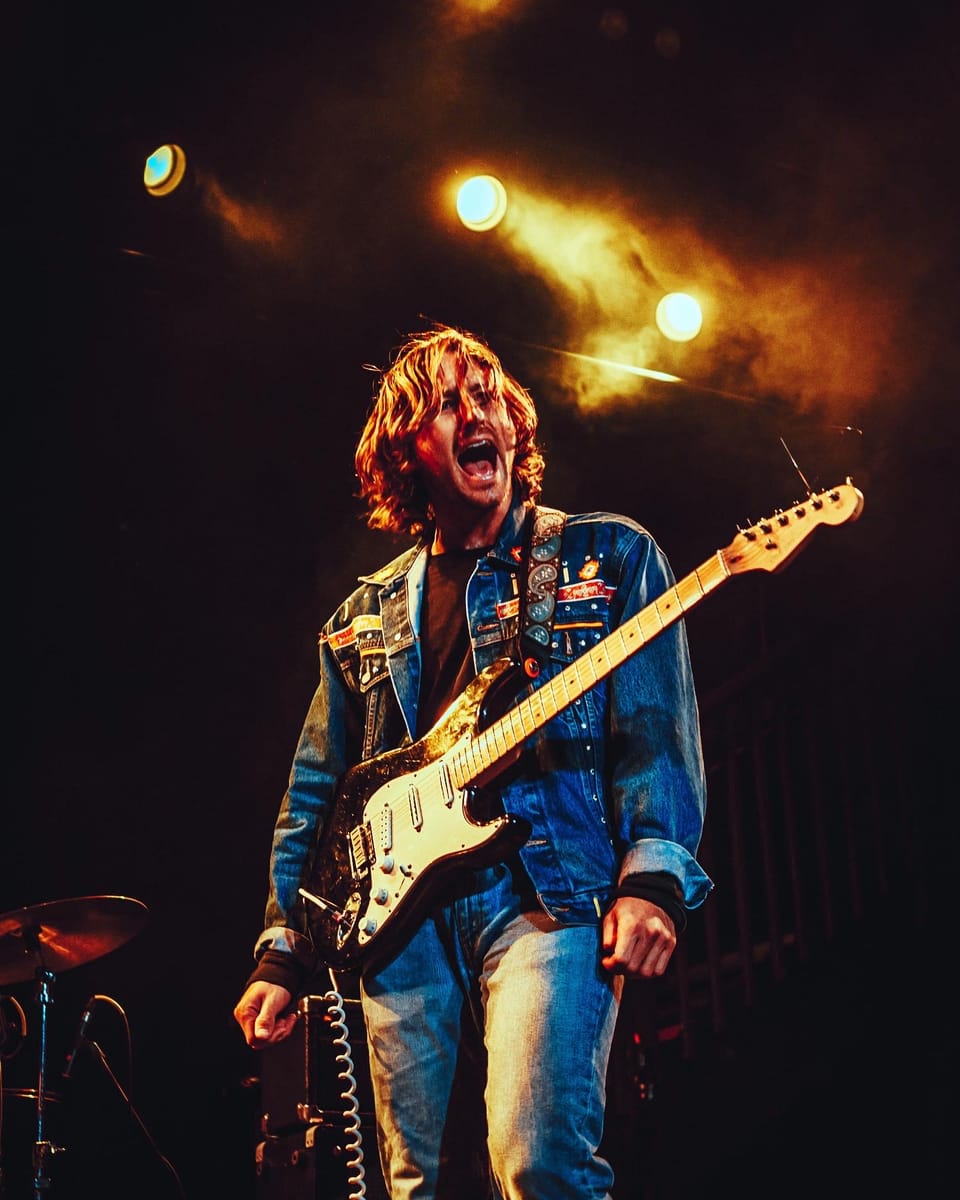 Easy Honey. A musician in a patched denim vest yells while playing a white Stratocaster in smoky light.