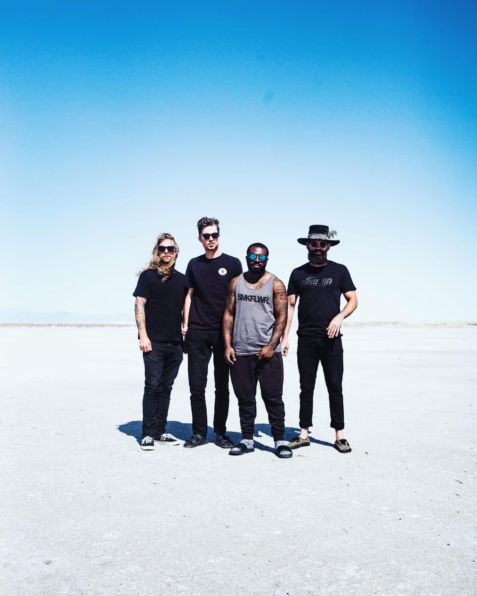 Of Good Nature. Four men stand in a row on a vast white salt flat under a pale blue sky.