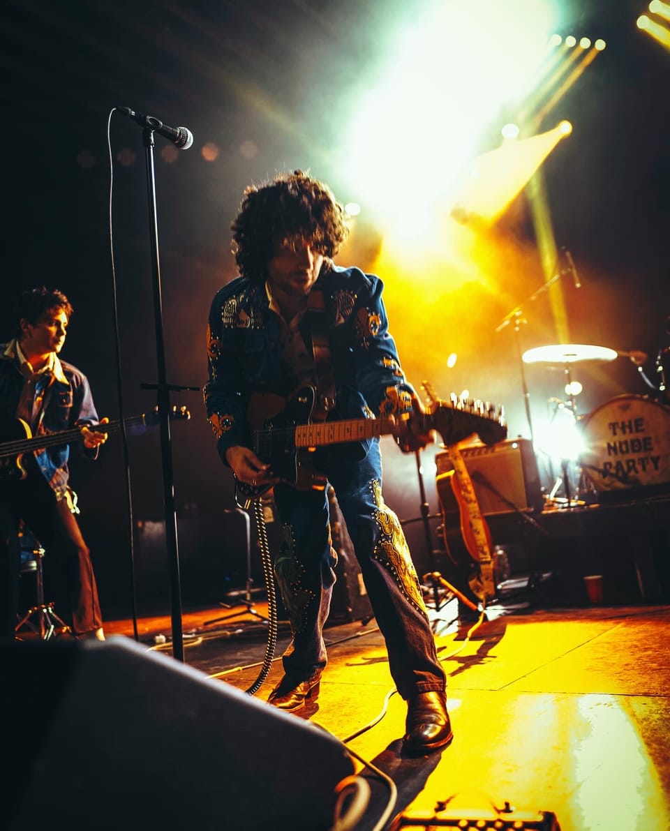 The Nude Party. A guitarist in a patterned jacket plays under a blaze of white and yellow light, another person beside him.