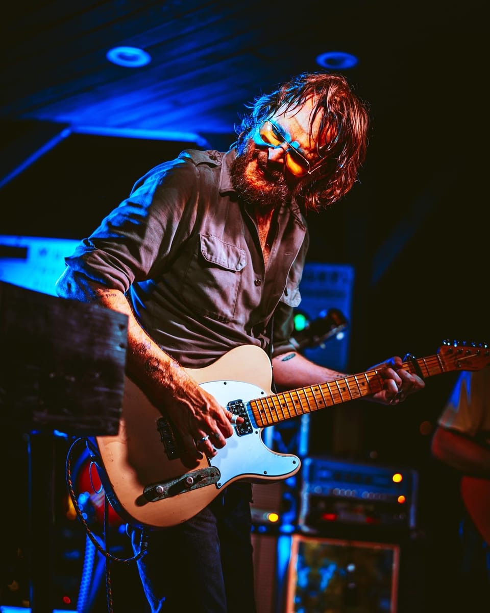 Hiss Golden Messenger. A bearded guitarist in a dark shirt leans over a Telecaster under blue stage light.