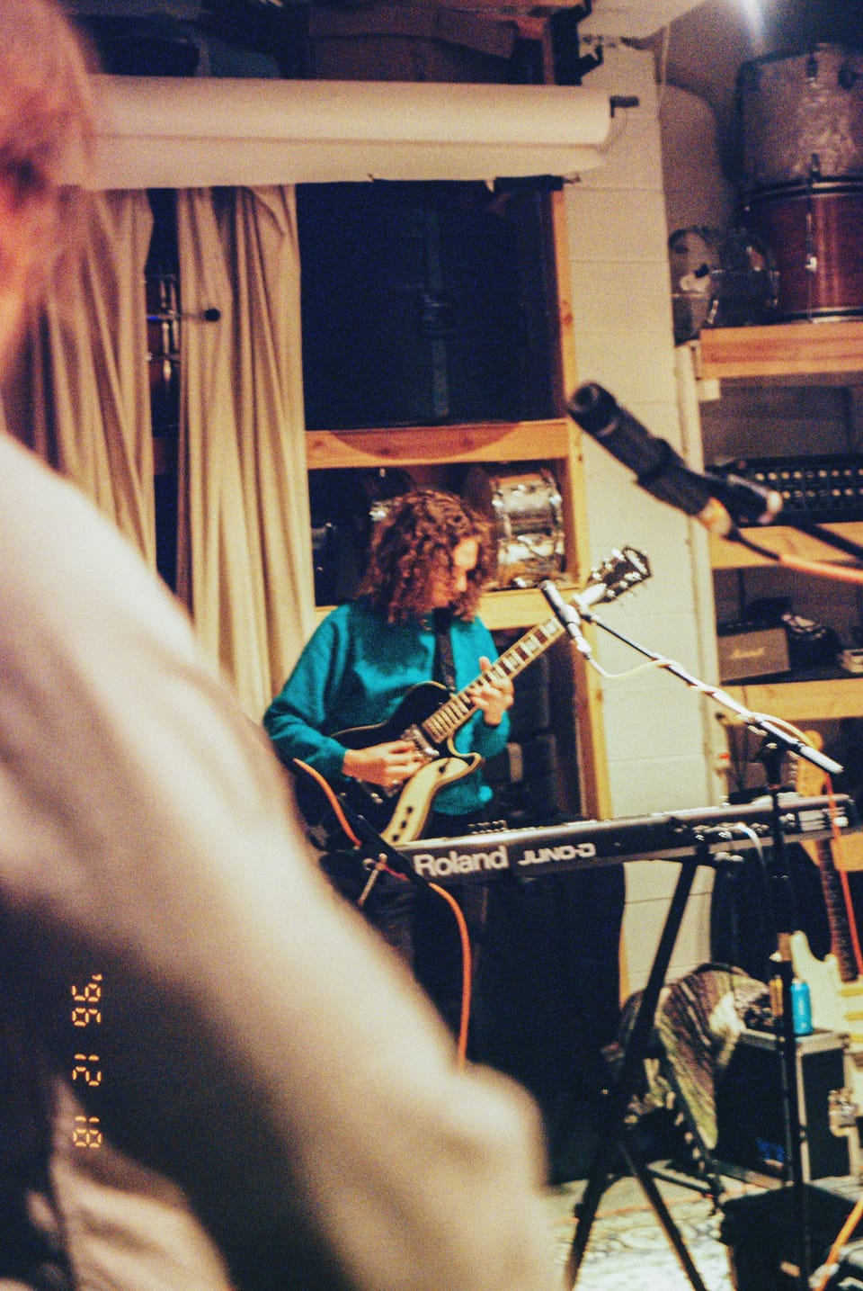 Mo Lowda and the Humble. A musician in a teal sweater plays guitar in a cluttered studio room with a keyboard in front.