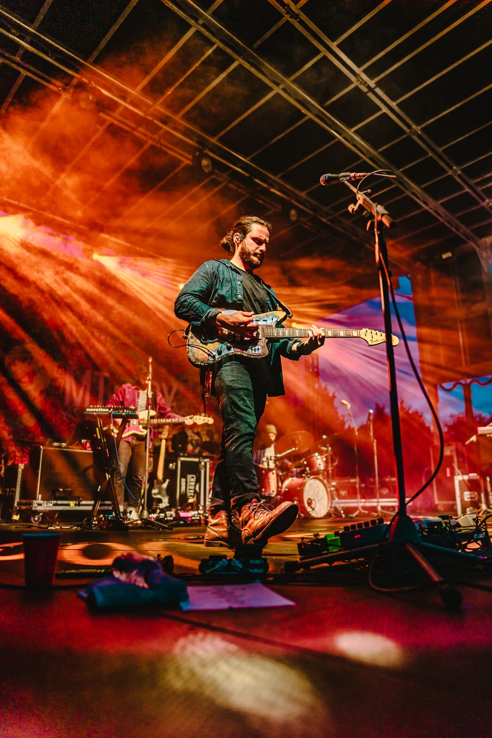 Mt. Joy. A guitarist plays on a big stage under orange haze and a steel truss.