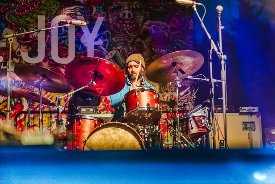 Mt. Joy. A drummer behind a red kit in front of a colourful mural backdrop with the word JOY.