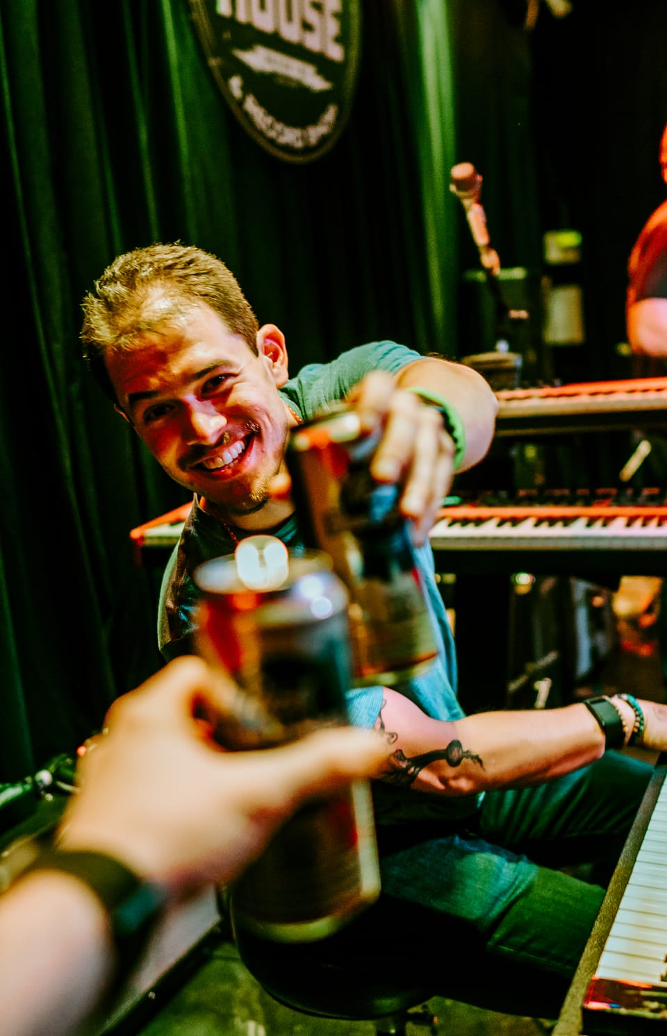 Joe May. A smiling musician at a keyboard clinks bottles with a hand reaching in from the foreground.