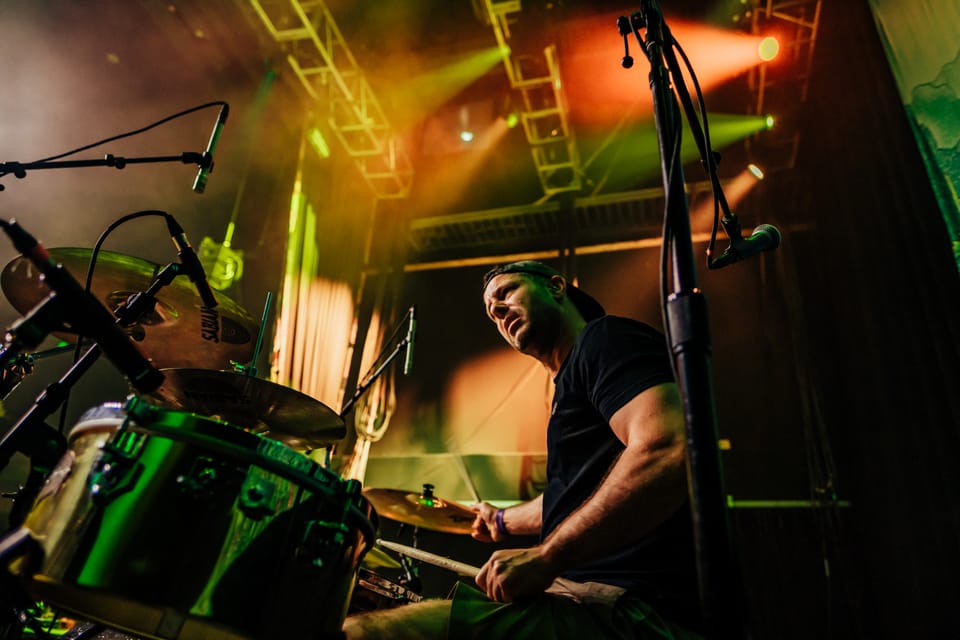 Badfish. A drummer plays under green and amber light, shot from a low angle through the kit.