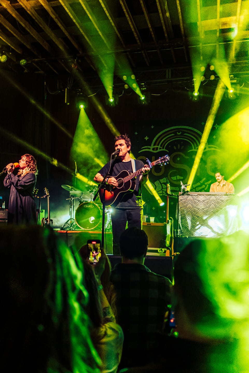 Susto. A guitarist plays on a stage washed in green light, a singer to his left, seen from the crowd.