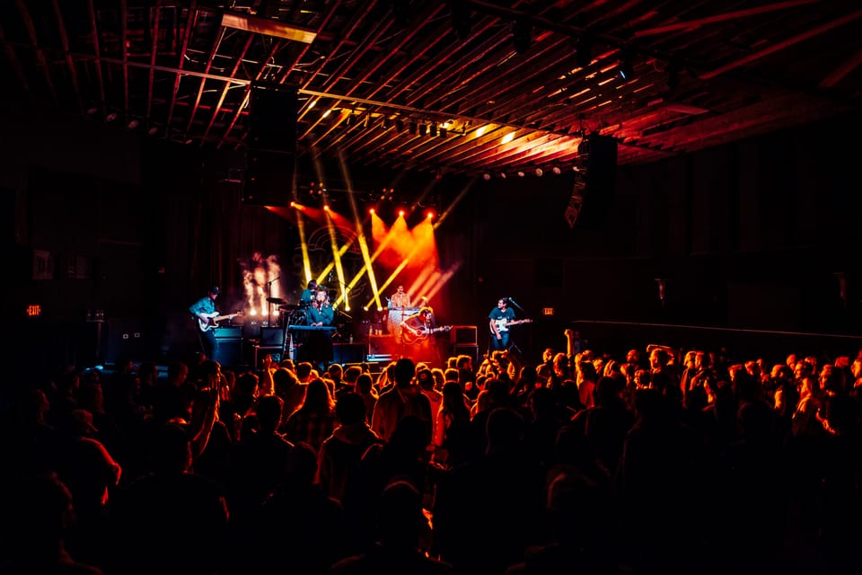 Susto. A wide shot of a band on a dark stage under red and orange beams, a crowd in front.