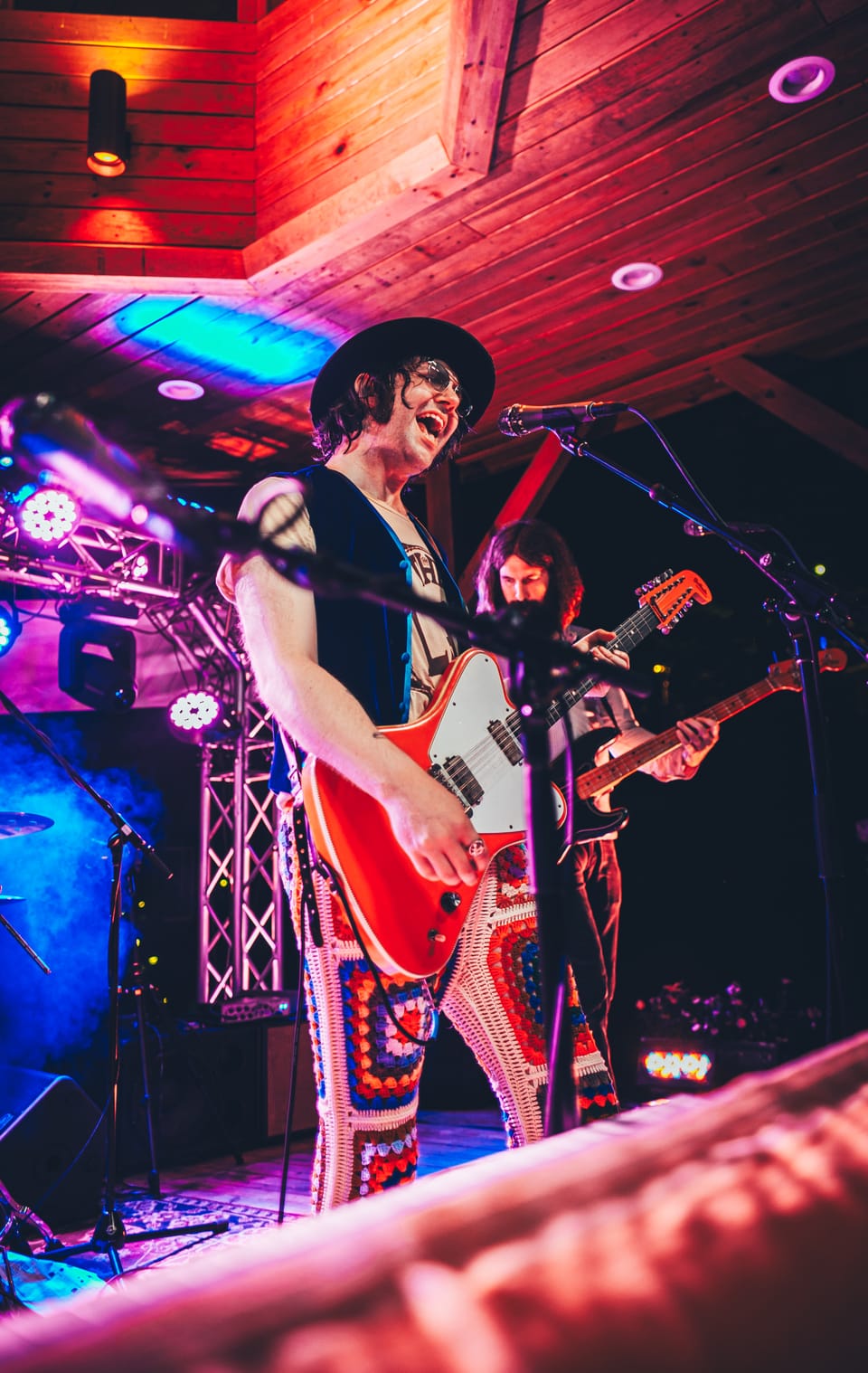 Aaron Lee Tasjan. A guitarist in a wide-brimmed hat and patterned trousers plays a red guitar on a wood-panelled stage.