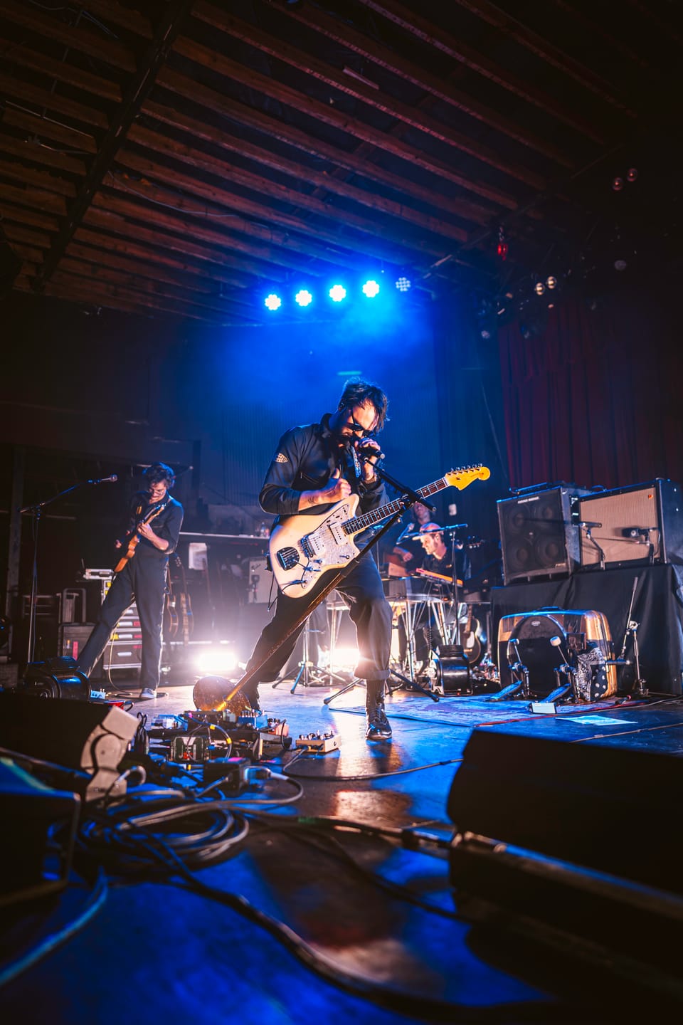 Shakey Graves. A guitarist on a dark stage under blue lights, seen from low at the front, with pedals and monitors in the foreground.