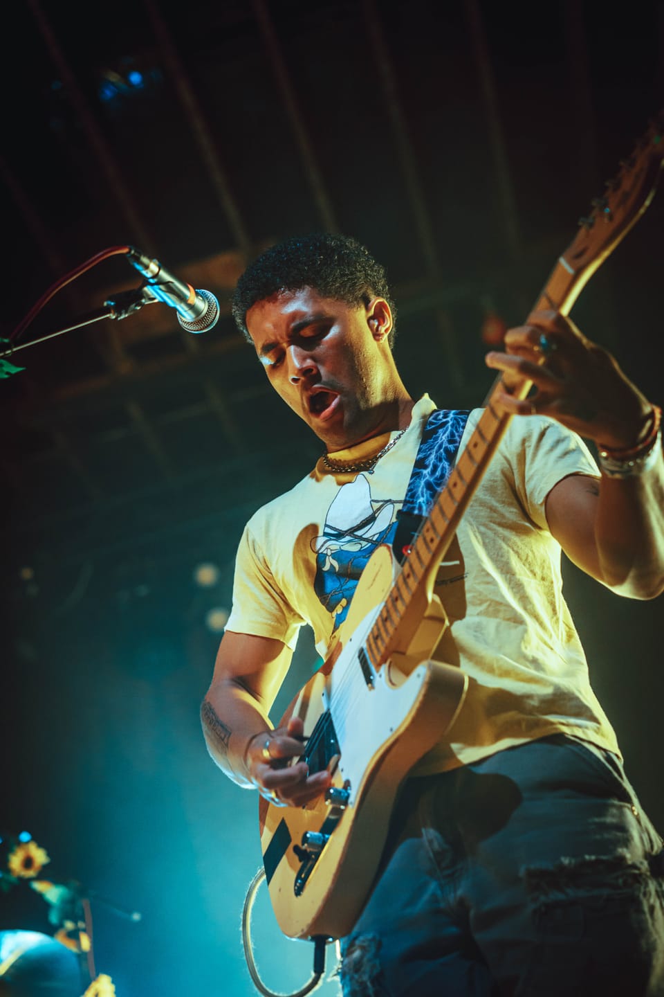 Briston Maroney. A guitarist in a yellow T-shirt plays under teal light, mouth open mid-song.