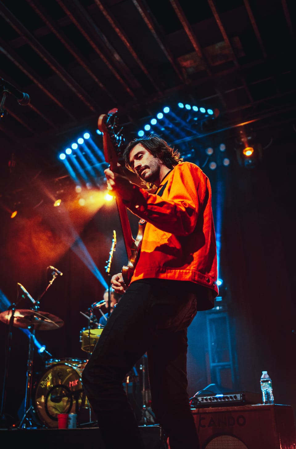 Houndmouth. A guitarist in an orange jacket plays on a stage lit by blue and orange lights, a drum kit behind him.