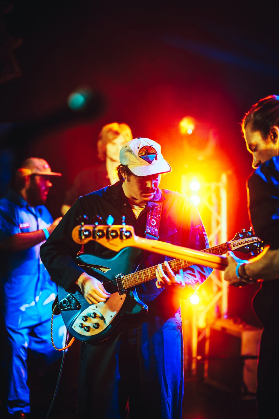 The New Creatures. A guitarist in a white cap plays a teal guitar among bandmates in red and orange light.