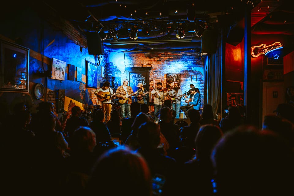 A band plays on a small club stage glowing blue and red, seen over the silhouettes of the crowd.