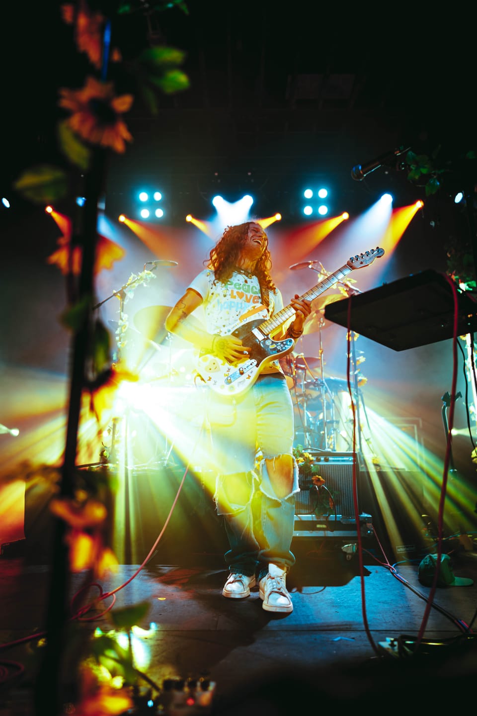 Briston Maroney. A guitarist backlit by a burst of golden stage lights, framed by hanging foliage.