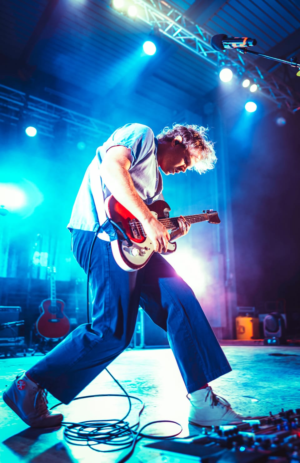 Colony House. A guitarist in a white shirt and blue trousers mid-stride on a stage washed in cyan light, cables at his feet.