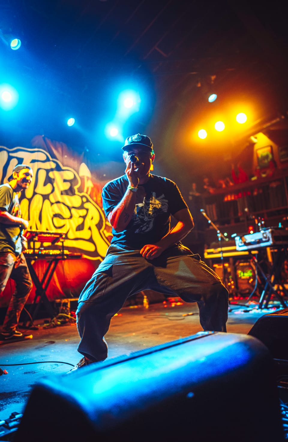 Little Stranger. A performer in a cap crouches on stage in front of a graffiti-style backdrop in blue and orange light.