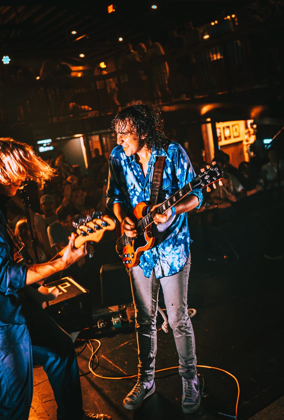 The Stews. A guitarist in a blue patterned shirt plays on a dim stage as a second player looks on.