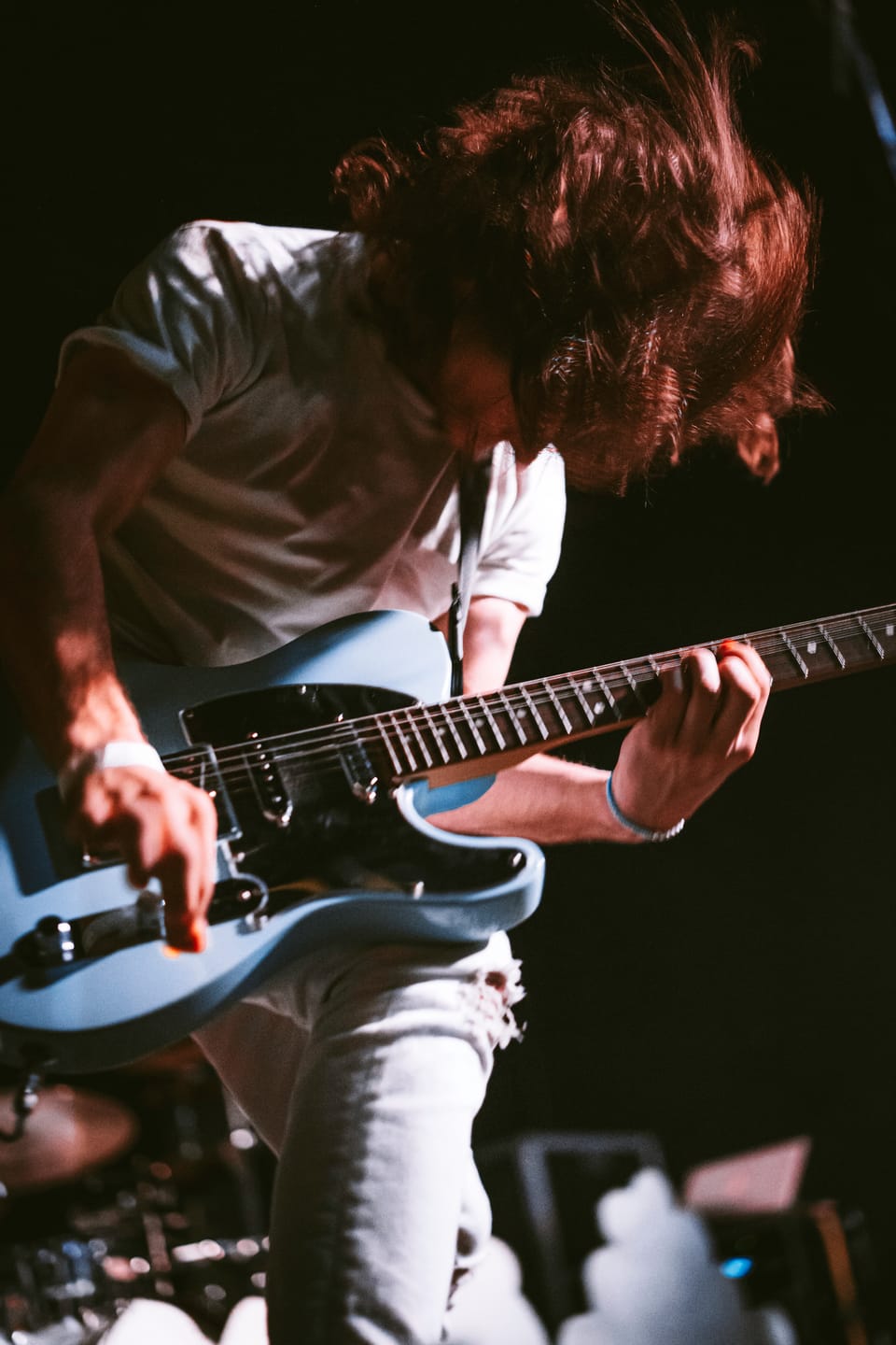 A guitarist bends over a pale blue guitar, his hair swinging forward.