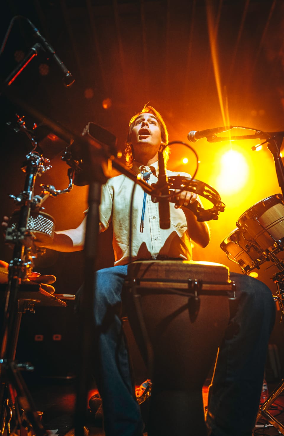 The Nude Party. A percussionist in a white shirt plays a hand drum under a glowing orange light.