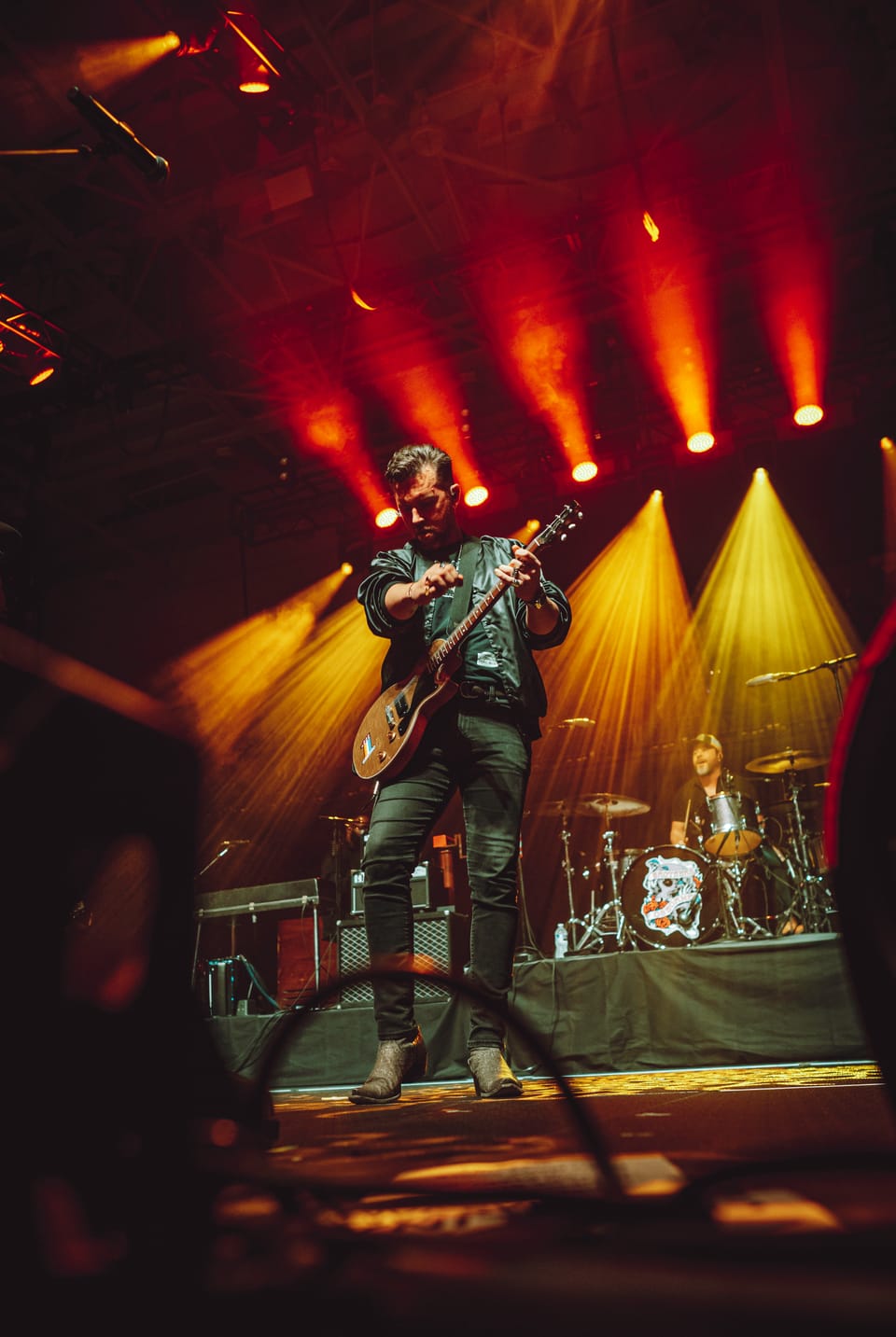 A guitarist plays on a stage lit by red and yellow beams, a drummer behind him.