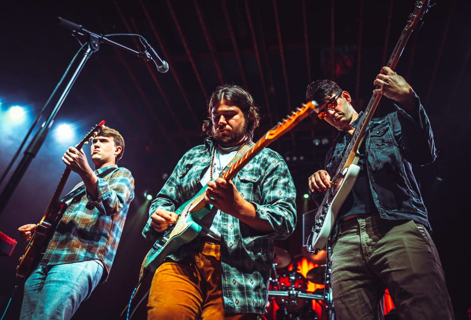 Joe May. Three guitarists in plaid shirts stand shoulder to shoulder on stage in blue light.