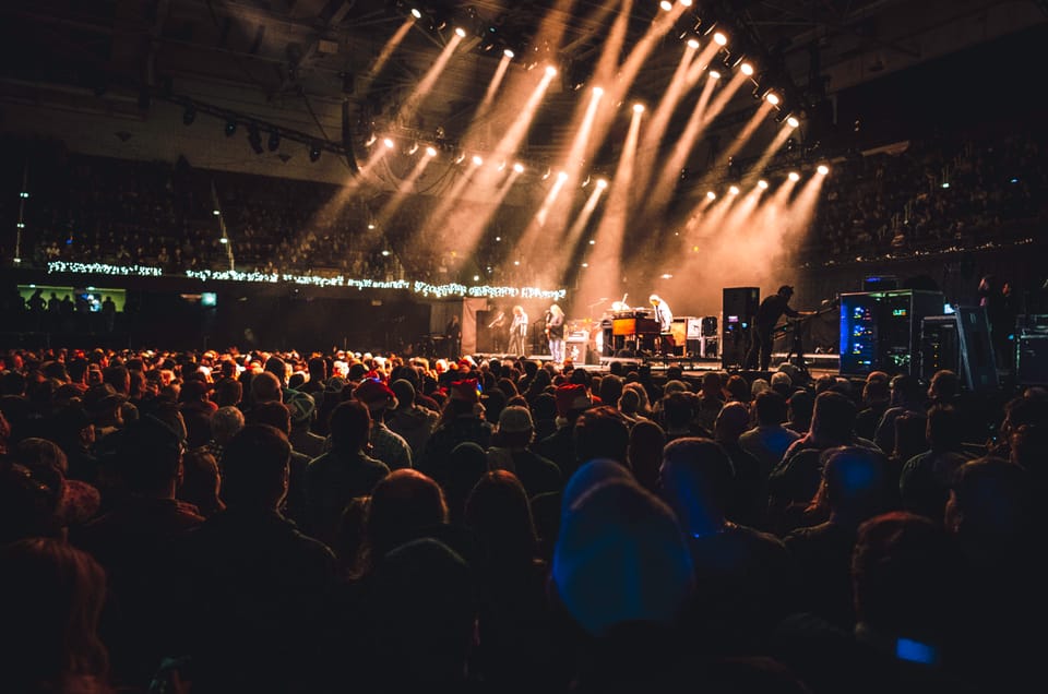 Warren Haynes. A wide view over a large crowd toward a distant stage lit with fanning white beams.