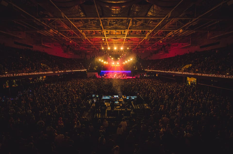 Warren Haynes. A packed arena seen from the back, a distant stage glowing under a coffered ceiling.