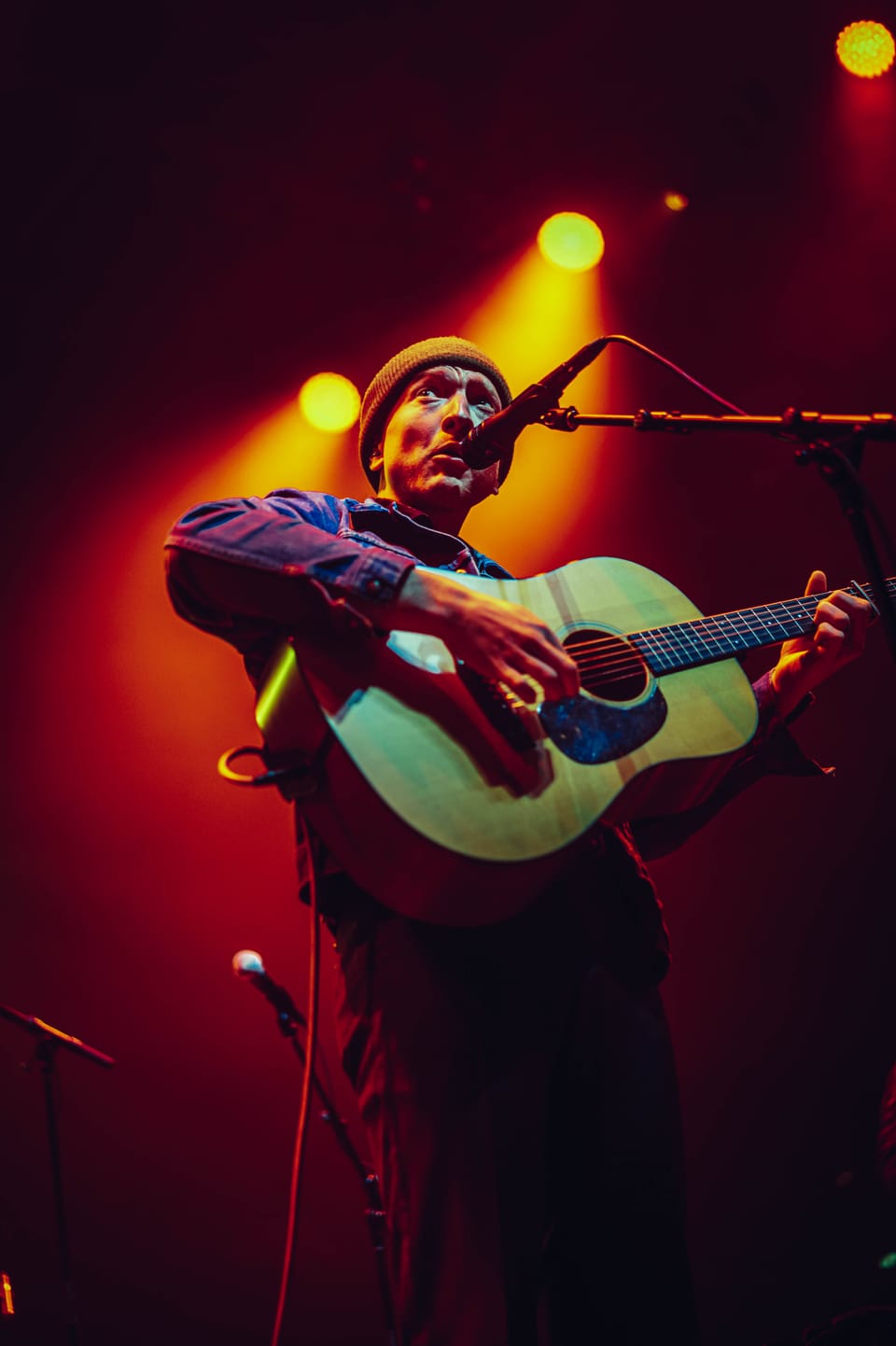 Tyler Childers. A musician in a cap plays an acoustic guitar, lit in red and yellow.