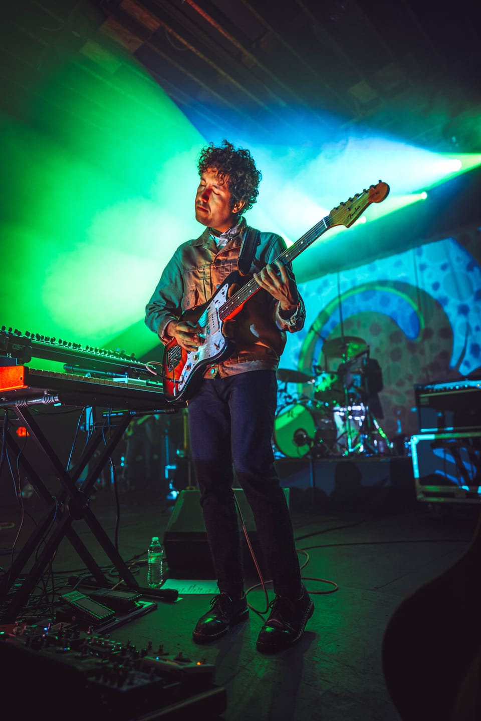Soccer Mommy. A guitarist in green light plays beside a keyboard, a patterned backdrop behind him.