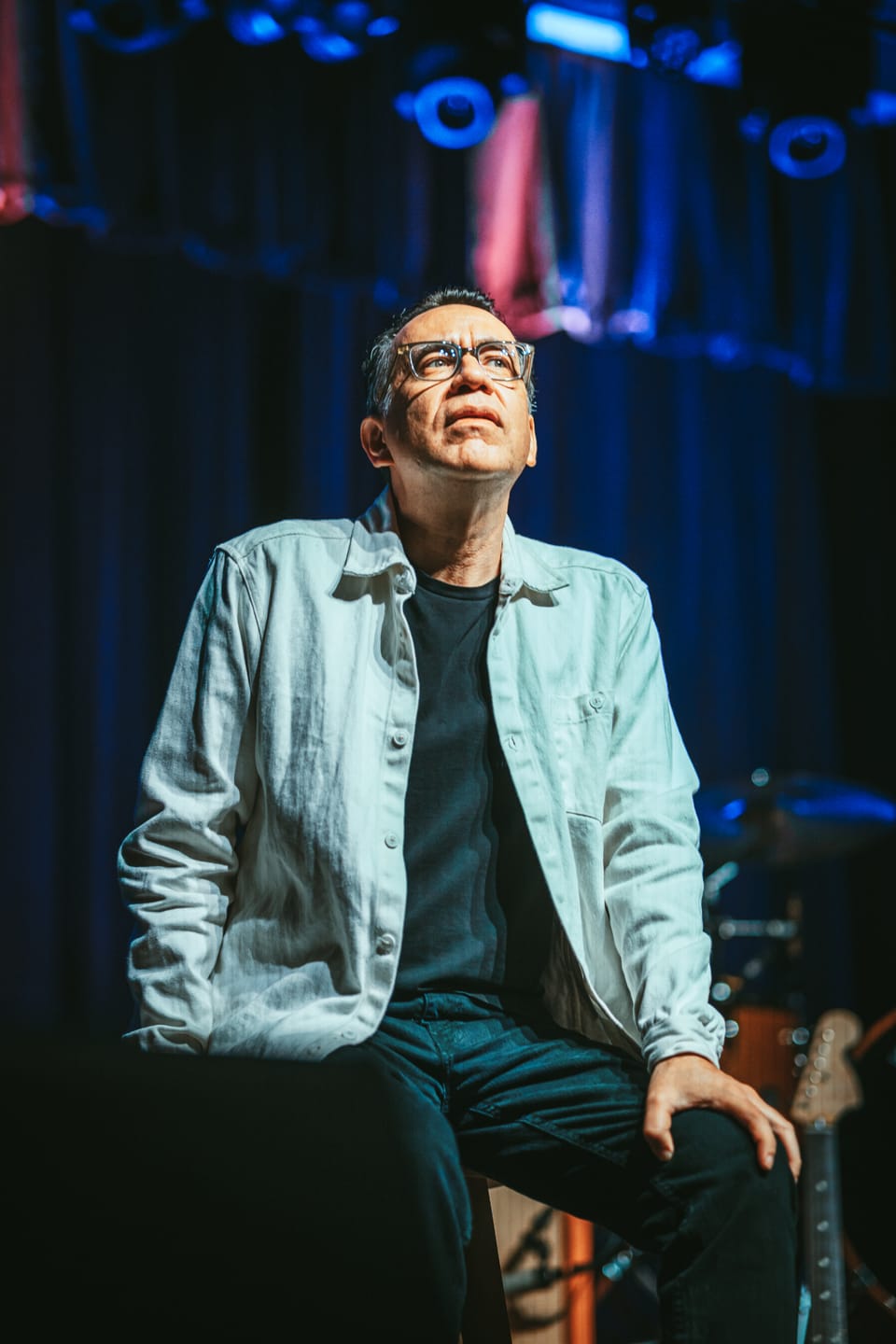 Fred Armisen. A man in glasses and a light jacket sits on a stool looking up, lit in blue.