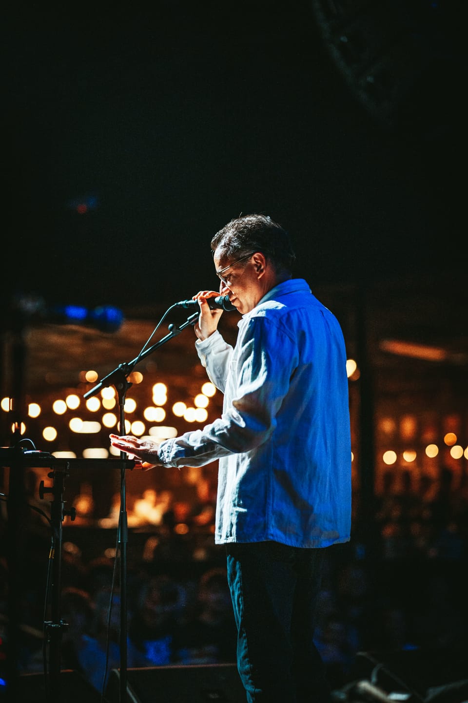 Fred Armisen. A man in a blue shirt sings at a microphone on a dark stage with string lights behind him.