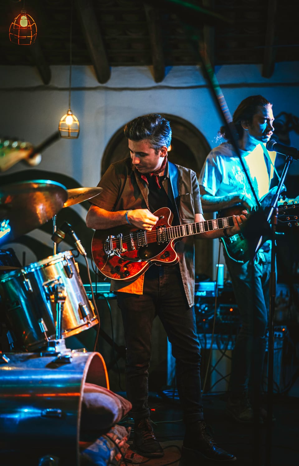 Young Mister. A guitarist plays a red hollow-body guitar beside a drum kit, a bandmate behind him.